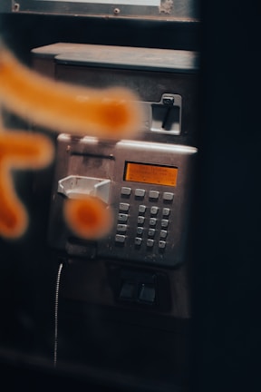 A close-up view of a metallic public payphone, featuring an alphanumeric keypad and an orange display screen. Some parts are out of focus due to a blurred orange pattern in the foreground.