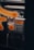 A close-up view of a metallic public payphone, featuring an alphanumeric keypad and an orange display screen. Some parts are out of focus due to a blurred orange pattern in the foreground.