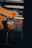 A close-up view of a metallic public payphone, featuring an alphanumeric keypad and an orange display screen. Some parts are out of focus due to a blurred orange pattern in the foreground.