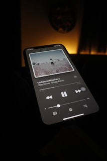 A smartphone screen is illuminated in a dimly lit room, displaying a music player application with a song titled 'Middle of Nowhere' by Vancouver Sleep Clinic. The screen shows album artwork depicting a serene outdoor scene.