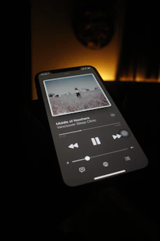 A smartphone screen is illuminated in a dimly lit room, displaying a music player application with a song titled 'Middle of Nowhere' by Vancouver Sleep Clinic. The screen shows album artwork depicting a serene outdoor scene.