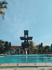 A swimming pool with clear blue water is surrounded by a metal fence. A tall, multi-level diving platform towers over the pool, with a backdrop of trees and buildings featuring red-tiled roofs. The sky is bright with some clouds and a palm tree partially visible to the side.