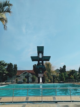A swimming pool with clear blue water is surrounded by a metal fence. A tall, multi-level diving platform towers over the pool, with a backdrop of trees and buildings featuring red-tiled roofs. The sky is bright with some clouds and a palm tree partially visible to the side.