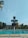 A swimming pool with clear blue water is surrounded by a metal fence. A tall, multi-level diving platform towers over the pool, with a backdrop of trees and buildings featuring red-tiled roofs. The sky is bright with some clouds and a palm tree partially visible to the side.
