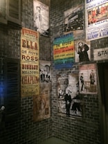 A collection of vintage theatrical posters displayed on a brick wall, featuring performances by various artists, including comedy and music acts. The posters exhibit a range of colors and artistic styles, reflecting an early 20th-century ambiance.