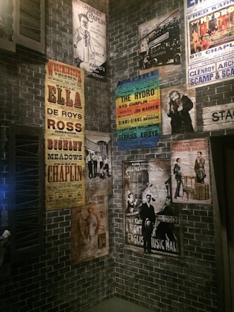 A collection of vintage theatrical posters displayed on a brick wall, featuring performances by various artists, including comedy and music acts. The posters exhibit a range of colors and artistic styles, reflecting an early 20th-century ambiance.