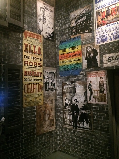 A collection of vintage theatrical posters displayed on a brick wall, featuring performances by various artists, including comedy and music acts. The posters exhibit a range of colors and artistic styles, reflecting an early 20th-century ambiance.