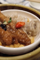 A close-up of a dish featuring a variety of ingredients. In the foreground, there is a piece of marinated or sauced meat garnished with chopped green onions and a red pepper slice. Adjacent to the meat, there are black beans and light brown lumps, possibly pieces of taro. The dish is presented in a white bowl set inside a bamboo steamer.