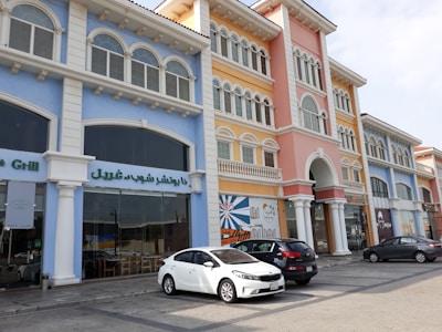 A row of colorful, two-story buildings featuring arches and columns. The facades are painted in pastel colors such as blue, pink, and yellow. Several cars are parked in front, and signage in Arabic is visible on the windows.