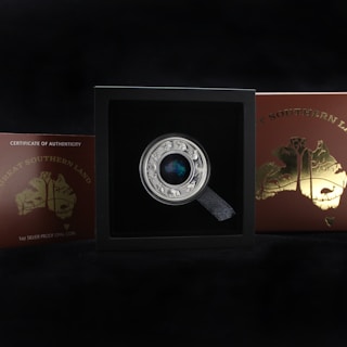 A collectible coin displayed in a black box with a label that reads 'Great Southern Land' and 'Certificate of Authenticity'. The coin has intricate designs with animals around the edge and a colorful central piece. The box and coin are set against a dark background.