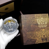 A hand wearing a white glove holds a decorative coin featuring a detailed design with wings, a face, and ornate elements. The background includes a box with intricate, golden Egyptian-themed artwork and the text 'AMUN RA DIVINE FACES OF THE SUN.' The scene suggests an ancient, mystical theme.