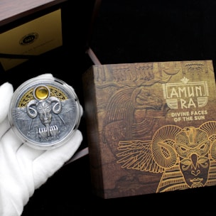 A hand wearing a white glove holds a decorative coin featuring a detailed design with wings, a face, and ornate elements. The background includes a box with intricate, golden Egyptian-themed artwork and the text 'AMUN RA DIVINE FACES OF THE SUN.' The scene suggests an ancient, mystical theme.