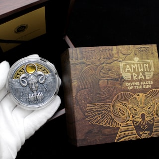 A hand wearing a white glove holds a decorative coin featuring a detailed design with wings, a face, and ornate elements. The background includes a box with intricate, golden Egyptian-themed artwork and the text 'AMUN RA DIVINE FACES OF THE SUN.' The scene suggests an ancient, mystical theme.
