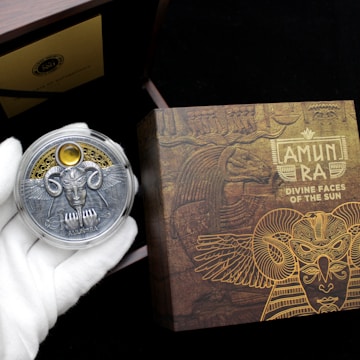 A hand wearing a white glove holds a decorative coin featuring a detailed design with wings, a face, and ornate elements. The background includes a box with intricate, golden Egyptian-themed artwork and the text 'AMUN RA DIVINE FACES OF THE SUN.' The scene suggests an ancient, mystical theme.