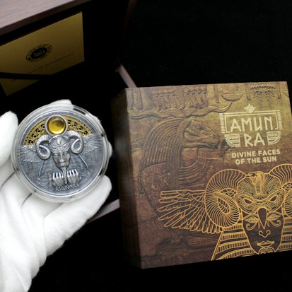 A hand wearing a white glove holds a decorative coin featuring a detailed design with wings, a face, and ornate elements. The background includes a box with intricate, golden Egyptian-themed artwork and the text 'AMUN RA DIVINE FACES OF THE SUN.' The scene suggests an ancient, mystical theme.