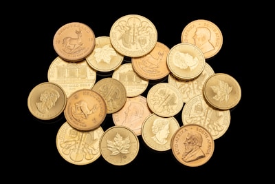 A collection of various gold coins featuring different designs, including portraits, animals, and emblems. The coins are arranged in a scattered manner on a black background, highlighting their shiny surfaces and intricate details.