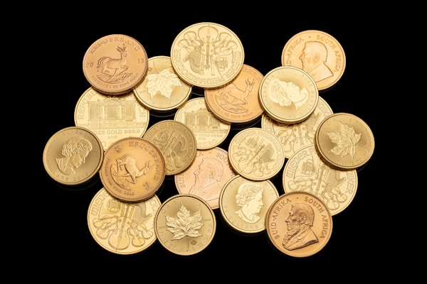A collection of various gold coins featuring different designs, including portraits, animals, and emblems. The coins are arranged in a scattered manner on a black background, highlighting their shiny surfaces and intricate details.