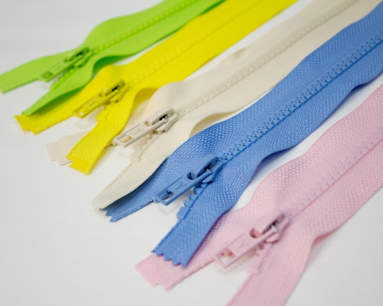 Multiple zippers in various pastel colors, including green, yellow, white, blue, and pink, are lined up next to each other on a white surface.