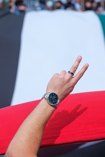A hand wearing a watch and a ring is making a peace sign gesture in front of a large fabric with colors resembling a flag. The background shows out-of-focus people.