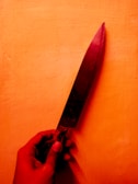 A hand is holding a large kitchen knife with a dark blade, set against a vibrant orange background. The lighting casts a warm tone, with shadows accentuating the details of the hand and knife.