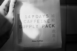 A hand is holding a package labeled with '14 Days Caffeine Supply Pack' and 'Akau Coffee' with some additional text in another language on the side. The package is captured in black and white, highlighting shadows and lighting.