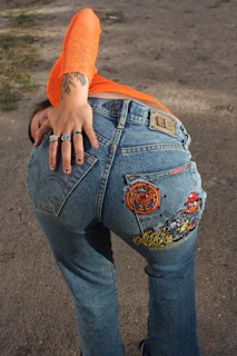 A person is wearing blue jeans with a cartoon graphic design on the back pocket. They have a tattoo on their hand and are wearing multiple rings. The person is also dressed in an orange mesh top, and the scene seems to be outdoors on an unpaved path.