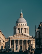 A neoclassical building with a large dome and Corinthian columns stands prominently in the background. Its facade features intricate architectural carvings and a French flag is visible. In the foreground, a street lined with cars and buses leads to the building, framed by buildings on both sides.