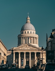 A neoclassical building with a large dome and Corinthian columns stands prominently in the background. Its facade features intricate architectural carvings and a French flag is visible. In the foreground, a street lined with cars and buses leads to the building, framed by buildings on both sides.