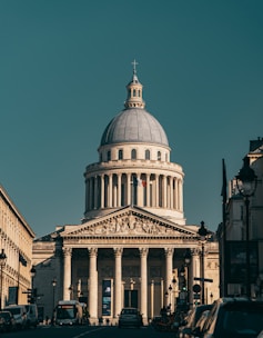 A neoclassical building with a large dome and Corinthian columns stands prominently in the background. Its facade features intricate architectural carvings and a French flag is visible. In the foreground, a street lined with cars and buses leads to the building, framed by buildings on both sides.