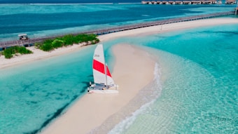 A catamaran with a red and white sail is parked on a pristine sandy beach surrounded by clear turquoise waters. A wooden bridge runs parallel to the beach, and small structures can be seen in the distance.