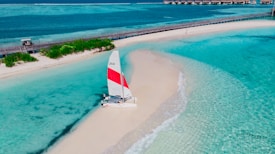 A catamaran with a red and white sail is parked on a pristine sandy beach surrounded by clear turquoise waters. A wooden bridge runs parallel to the beach, and small structures can be seen in the distance.