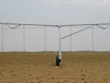 A large agricultural irrigation system is visible in a barren, flat field under an overcast sky. The system includes a long metallic structure with hoses hanging down from it, supported by a wheel.