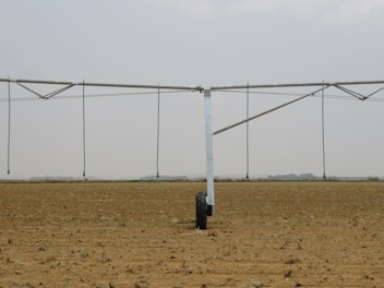 A large agricultural irrigation system is visible in a barren, flat field under an overcast sky. The system includes a long metallic structure with hoses hanging down from it, supported by a wheel.