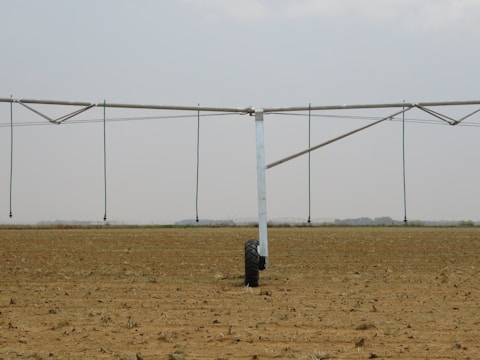 A large agricultural irrigation system is visible in a barren, flat field under an overcast sky. The system includes a long metallic structure with hoses hanging down from it, supported by a wheel.