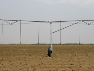 A large agricultural irrigation system is visible in a barren, flat field under an overcast sky. The system includes a long metallic structure with hoses hanging down from it, supported by a wheel.