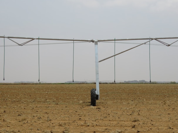 A large agricultural irrigation system is visible in a barren, flat field under an overcast sky. The system includes a long metallic structure with hoses hanging down from it, supported by a wheel.