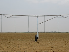 A large agricultural irrigation system is visible in a barren, flat field under an overcast sky. The system includes a long metallic structure with hoses hanging down from it, supported by a wheel.