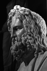 A detailed marble sculpture depicting the head and upper torso of a bearded man with curly hair. The craftsmanship highlights intricate details in the hair and beard, showcasing the skill of the sculptor. Shadows cast by the lighting add depth and emphasize the texture of the stone.