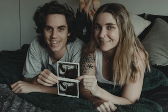 A couple is lying on a bed, smiling at the camera, while holding ultrasound pictures. The man has curly hair and wears a gray t-shirt. The woman has long blonde hair, a nose ring, and a tattoo visible on her arm. They look happy and relaxed, surrounded by pillows and blankets.