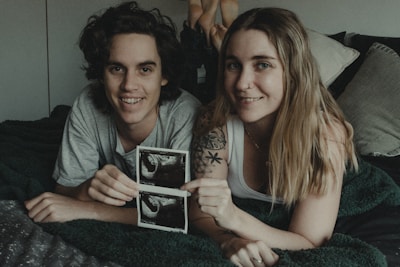 A couple is lying on a bed, smiling at the camera, while holding ultrasound pictures. The man has curly hair and wears a gray t-shirt. The woman has long blonde hair, a nose ring, and a tattoo visible on her arm. They look happy and relaxed, surrounded by pillows and blankets.