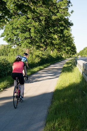 Two cyclists ride along a paved path surrounded by lush green trees and grass on a sunny day. The sun casts long shadows, creating a serene and inviting atmosphere. The cyclists are wearing helmets and sporty attire, cycling in a peaceful natural setting.