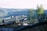 A viewing platform extends into a scenic landscape with people enjoying the view. The platform is elevated and supported by beams, offering a panoramic outlook over a lush, forested area and a body of water. The hills and trees in the background create a serene natural atmosphere.
