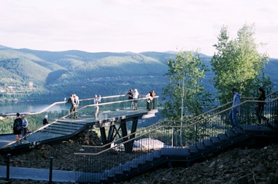 A viewing platform extends into a scenic landscape with people enjoying the view. The platform is elevated and supported by beams, offering a panoramic outlook over a lush, forested area and a body of water. The hills and trees in the background create a serene natural atmosphere.