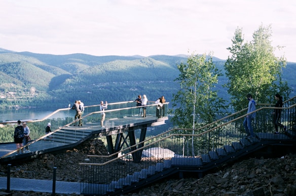 A viewing platform extends into a scenic landscape with people enjoying the view. The platform is elevated and supported by beams, offering a panoramic outlook over a lush, forested area and a body of water. The hills and trees in the background create a serene natural atmosphere.