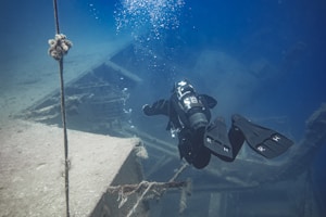 A scuba diver explores a sunken shipwreck underwater. The diver is equipped with full diving gear, including a tank and flippers, and is surrounded by the deep blue of the ocean. The shipwreck structure appears aged and covered in some marine growth.