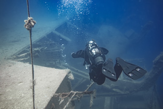 A scuba diver explores a sunken shipwreck underwater. The diver is equipped with full diving gear, including a tank and flippers, and is surrounded by the deep blue of the ocean. The shipwreck structure appears aged and covered in some marine growth.