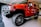 A bright red SUV with large off-road tires is parked indoors on a shiny black and gray floor. The vehicle has a robust front grille, side steps, and prominent side mirrors. The ceiling lighting reflects off the polished surface of the vehicle.