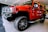 A bright red SUV with large off-road tires is parked indoors on a shiny black and gray floor. The vehicle has a robust front grille, side steps, and prominent side mirrors. The ceiling lighting reflects off the polished surface of the vehicle.