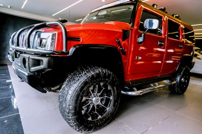 A bright red SUV with large off-road tires is parked indoors on a shiny black and gray floor. The vehicle has a robust front grille, side steps, and prominent side mirrors. The ceiling lighting reflects off the polished surface of the vehicle.