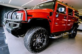 A bright red SUV with large off-road tires is parked indoors on a shiny black and gray floor. The vehicle has a robust front grille, side steps, and prominent side mirrors. The ceiling lighting reflects off the polished surface of the vehicle.